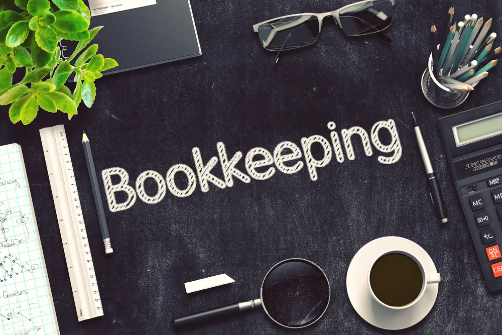 Bookkeeping Text on Black Irish Business Desk with Coffee in a Cup, Calculator, Pens in Tin. Ruler, Eyeglasses